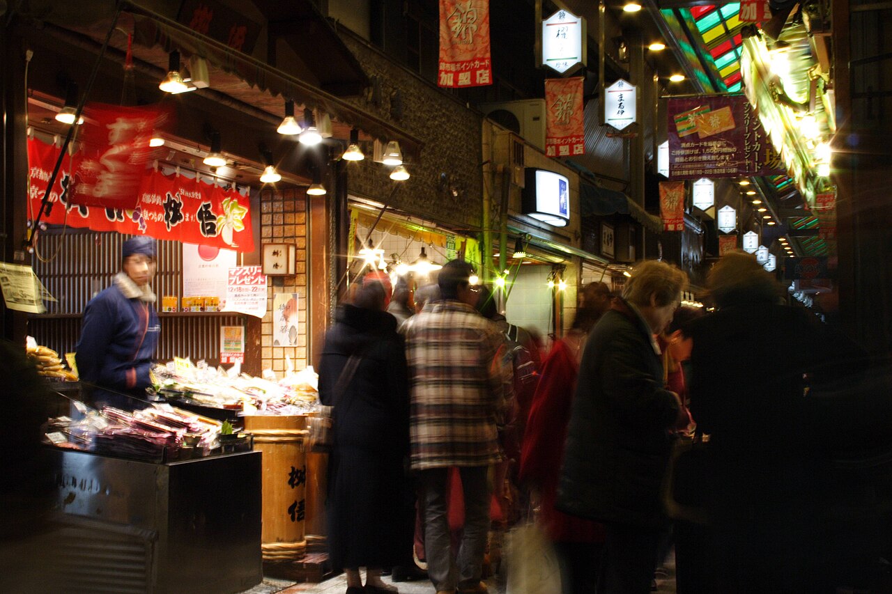 A bustling covered arcade of Nishiki Market in Kyoto, with various food stalls and shoppers.
