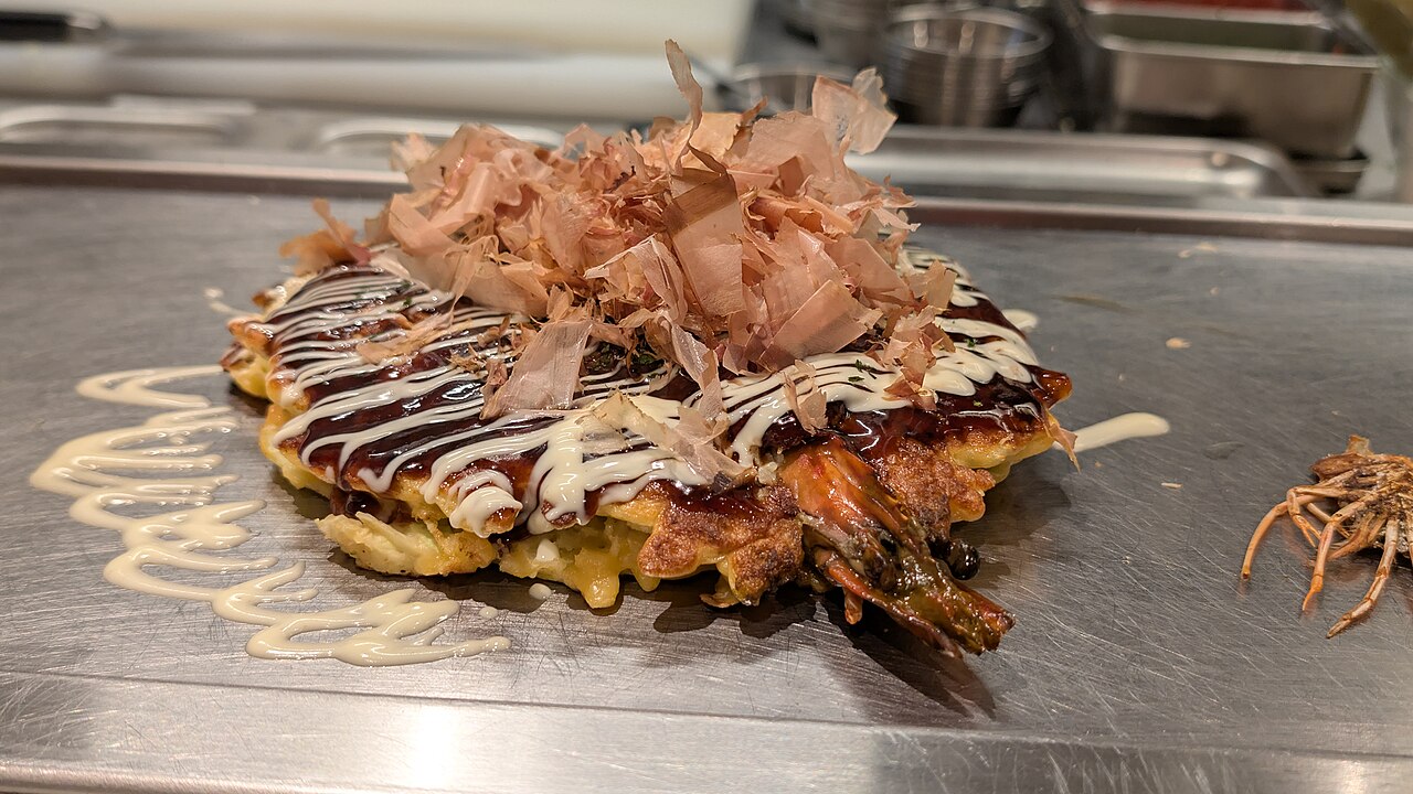 Osaka-style okonomiyaki cooking on a griddle, topped with sauce and mayonnaise